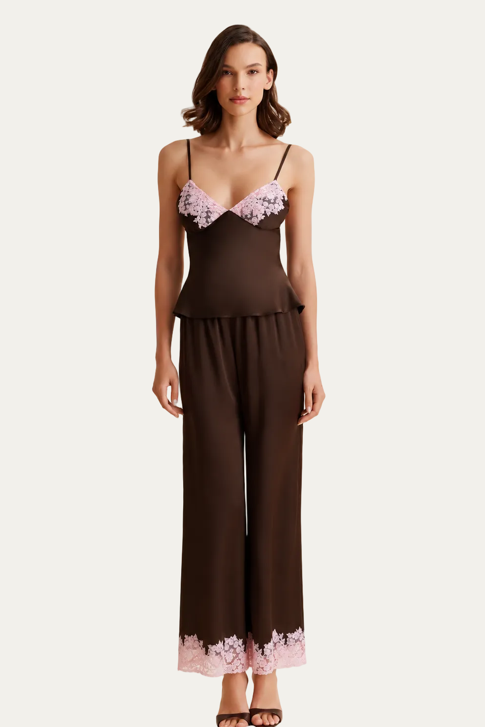 A woman models a two-piece satin pajama set consisting of a dark brown camisole top with light pink lace cups and matching wide-leg pants with a light pink lace hem, set against a draped cream fabric backdrop.