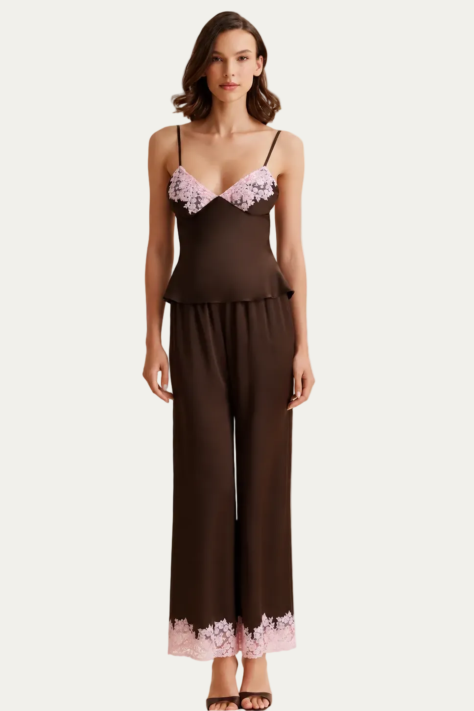 A woman models a two-piece satin pajama set consisting of a dark brown camisole top with light pink lace cups and matching wide-leg pants with a light pink lace hem, set against a draped cream fabric backdrop.