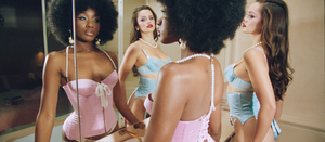 Two women looking in the mirror, one in a pink corset, one in a blue lingerie set