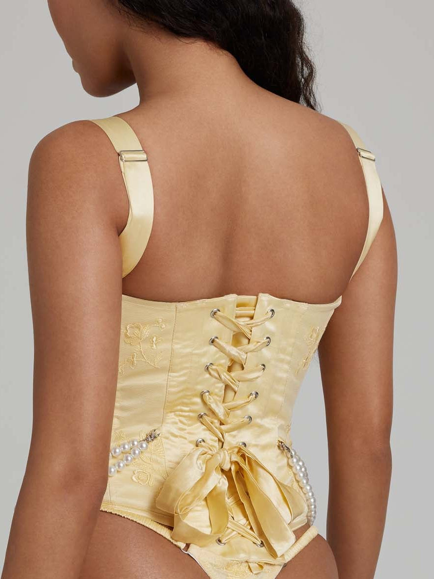 Woman wearing yellow corset with pearl detailing and busk closures and yellow thong with white bows from behind