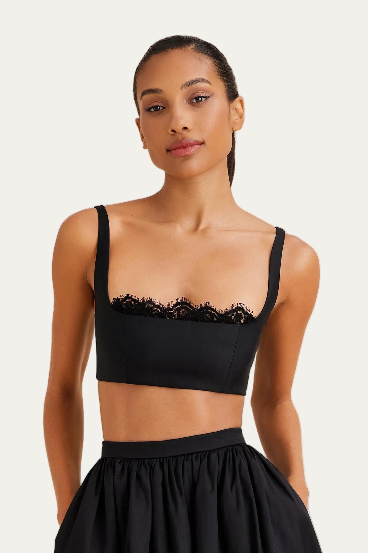 A model wearing a black two-piece outfit consisting of a cropped camisole top with lace trim and a matching high-waisted pleated midi skirt, paired with black strappy heels, against a light gray studio background.