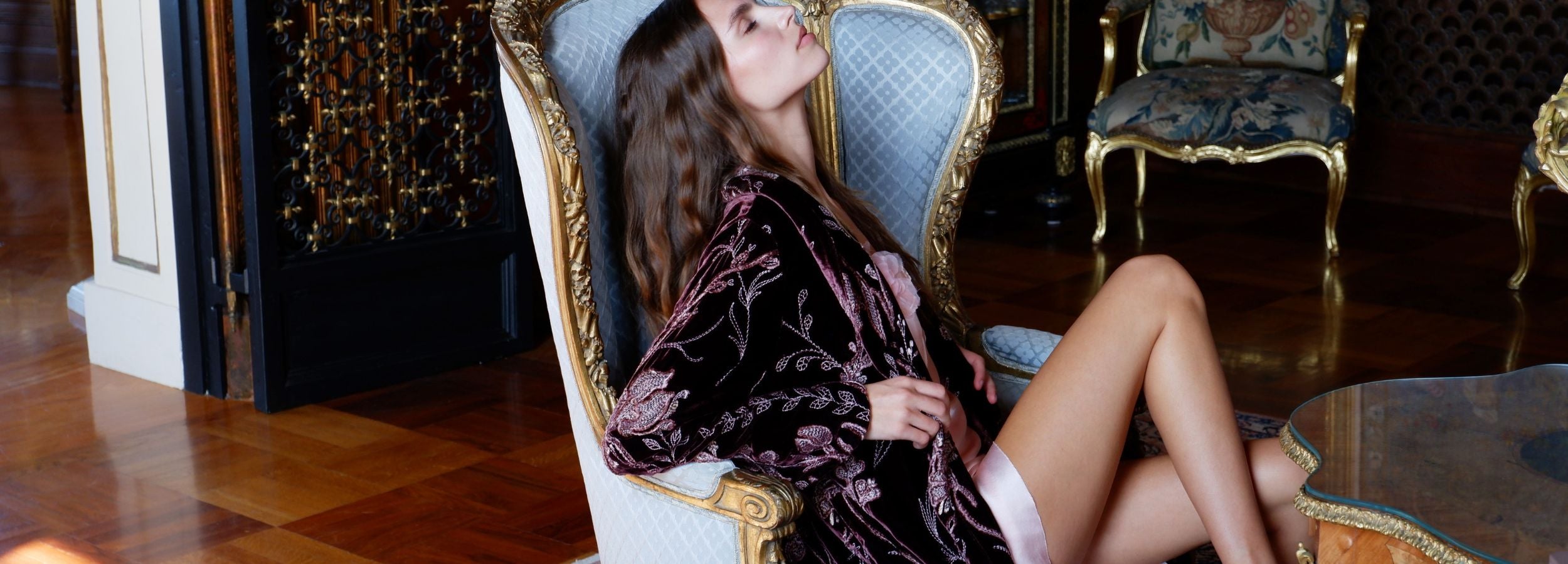 Woman in a luxurious robe sitting on an ornate chair in a opulent room.